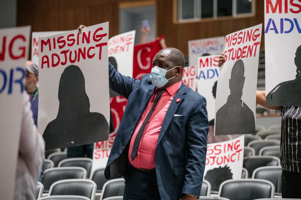 A PSC member in the audience at a CUNY Board of Trustees hearing May 8, 2023 at LaGuardia Community College holds a sign that reads, "Missing Adjunct," surrounded by other missing signs.